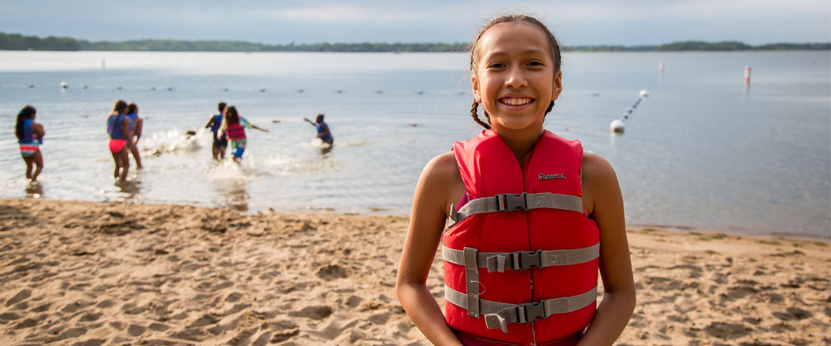 a girl in a life jacket smiling on the beach with kids playing in the water behind her.