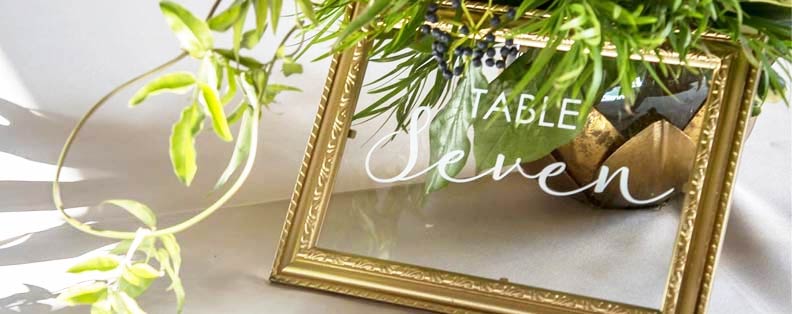 a gold frame with white lettering on the glass that says table seven. It's tucked under a plant.