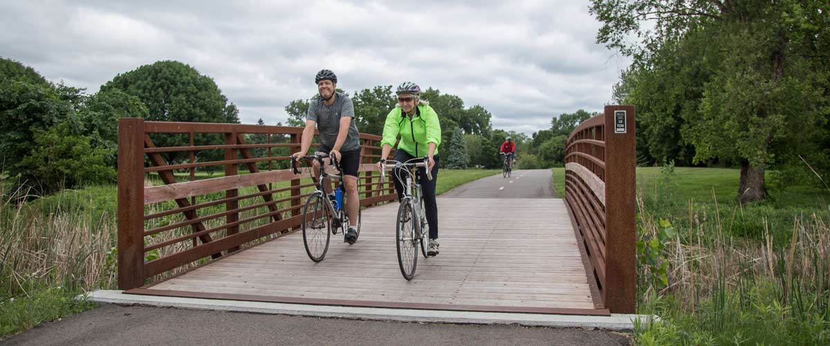 a man in a gray t-shirt and a woman in a neon green jacket ride their bikes over a wooden bridge on a bike path.