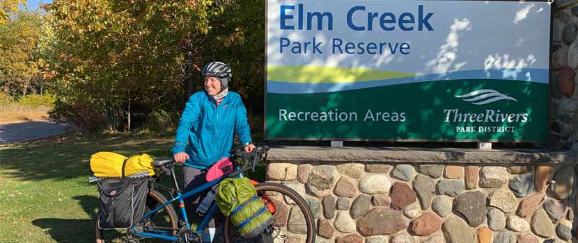A woman stands in front of the Elm Creek Park Reserve sign with a bike packed for camping.
