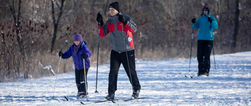 Three people cross-country ski on a snowy trail.