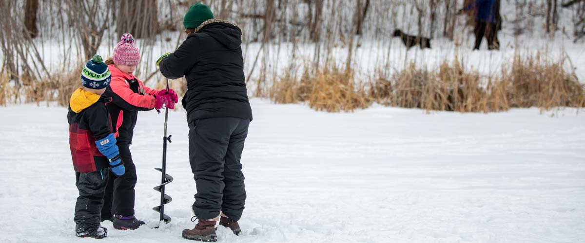 Two people drill into the ice for ice fishing while one child watches.