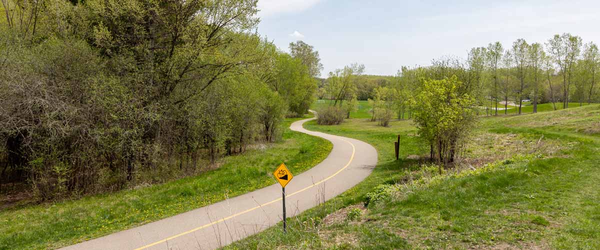 A paved trail weaves through lush green grasses and budding trees. A sign indicating a hill with a bike is close to the trail.
