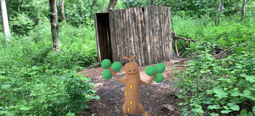 A tree-like video game character stands in front of a wooden sculpture at a park.
