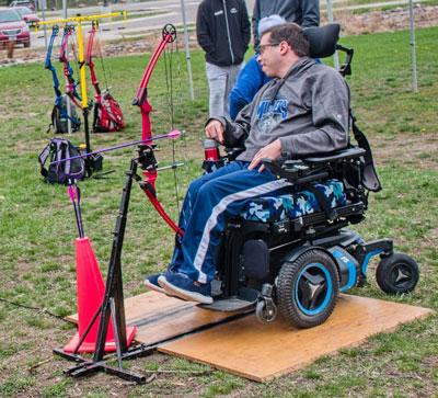 A bow stand with a bow is in front of a person in a wheelchair at the archery range.