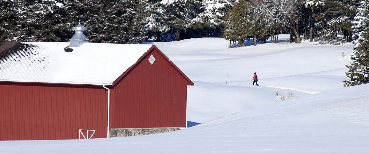 A cross-country skier skis by a red building in the snow.