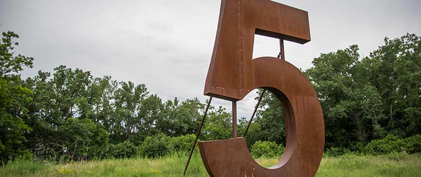 a giant metal number 5 sculpture outside with trees in the background.