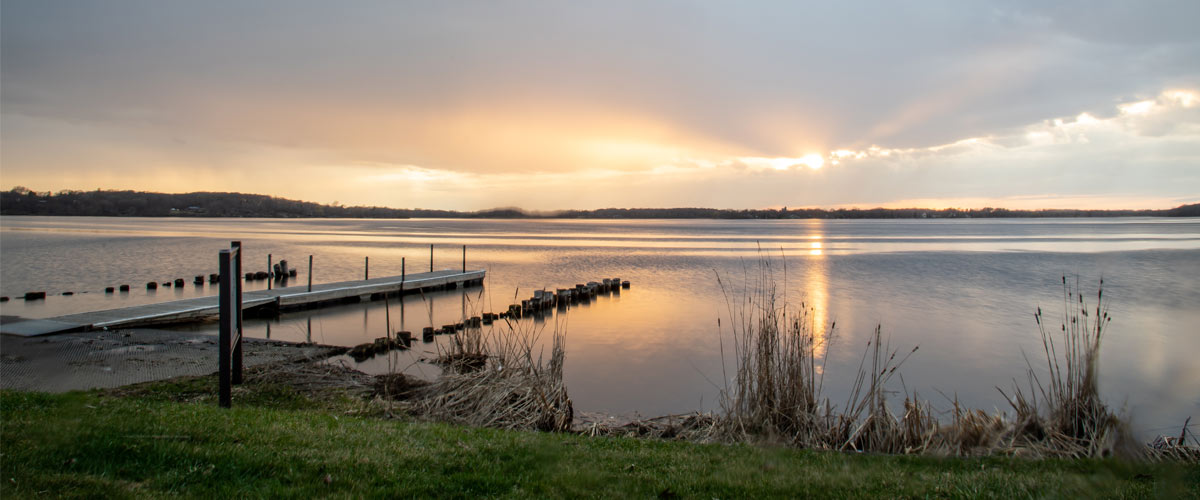 The sun sets over a https://www.threeriversparks.org/sites/default/files/2019-04/Baker%20Sunset%202019.jpglake. The edge of the water with a dock are in the foreground.