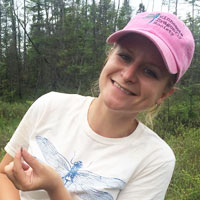 a woman in pink baseball cap and white shirt holds a dragonfly and smiles at the camera.