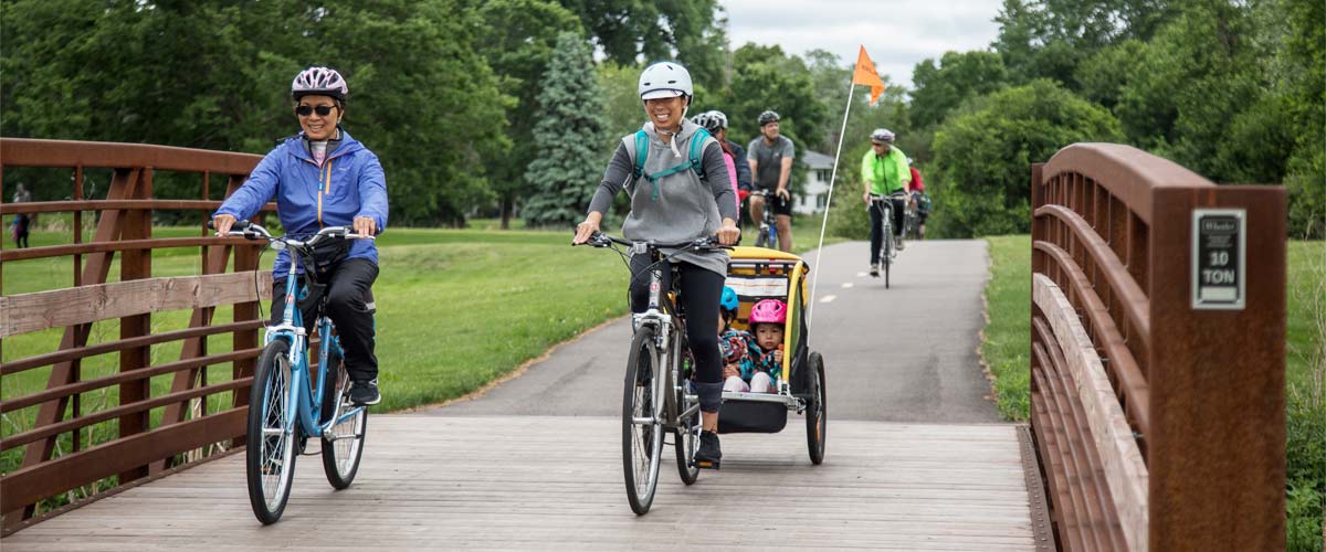 Two women ride their bikes over a bridge on a bike path. One is pulling a trailer with children in it.