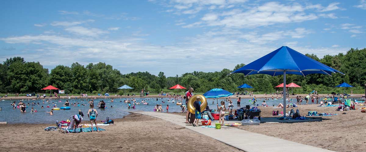 A swimming pond surrounded by sand and trees. The skies are blue and many people are in the water or laying on towels in the sand. Some are under large red or blue umbrellas in the sand.
