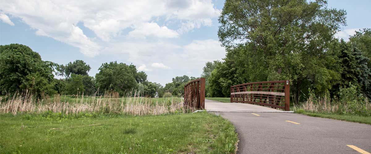 a paved trail that goes over a small bridge. The trail is surrounded by open grass and trees in some areas. The sky is blue with large clouds.