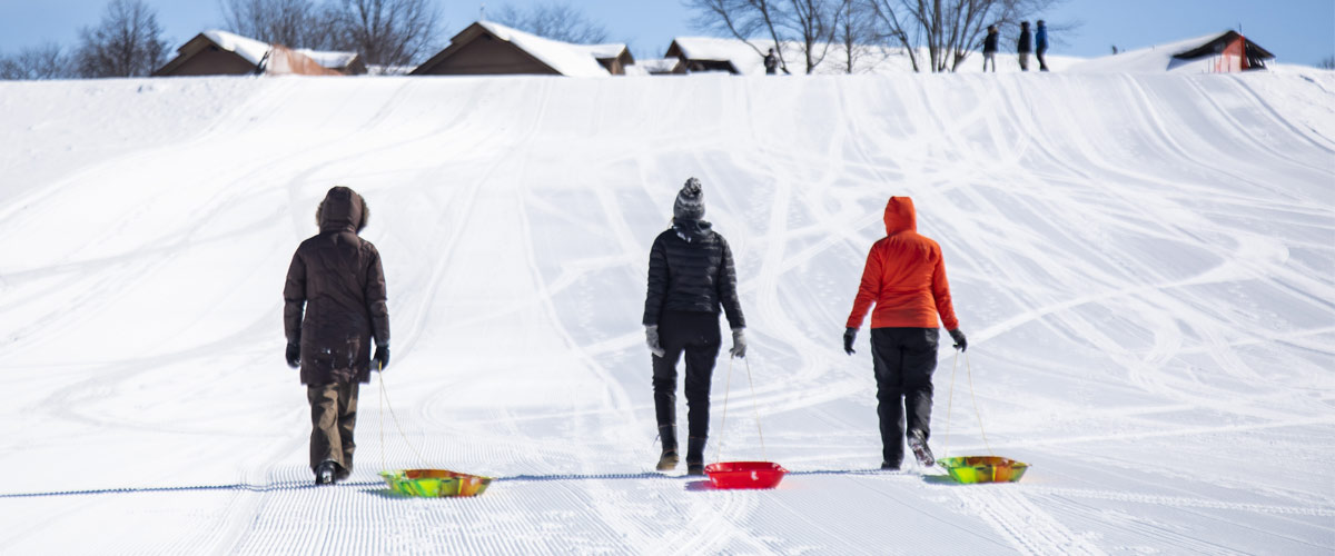 Three women walk up a sledding hill dragging colorful plastic sleds behind them.