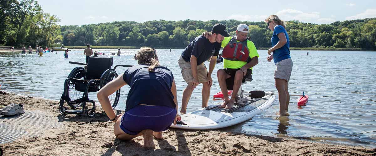 a man is helped from his wheelchair onto an adapted stand-up paddleboard.