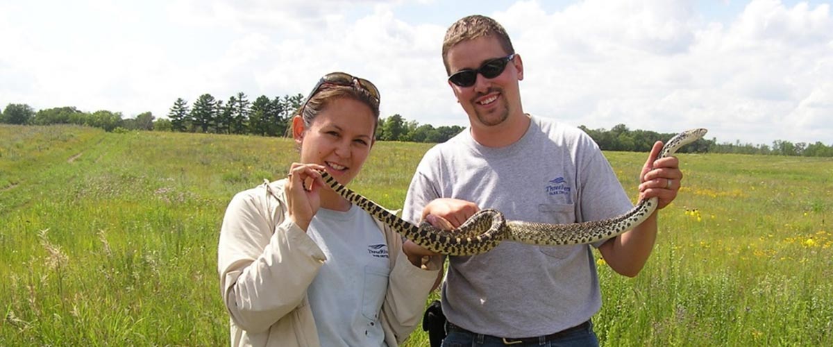 A woman and a man standing in a field hold up a long snake.