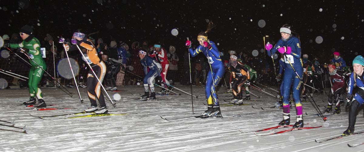 High school girls compete in a cross-country skiing race at night while it snows.