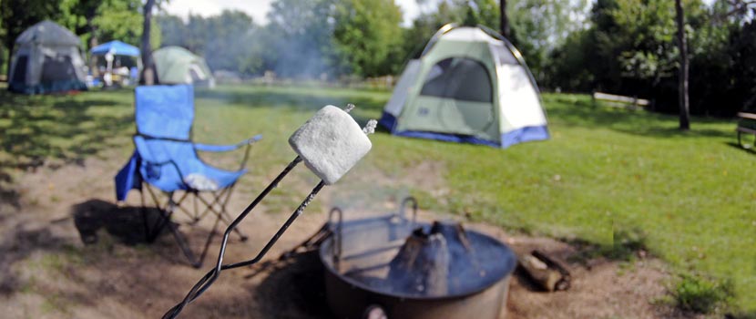 A marshmallow on a roasting stick in front of a campsite with a tent, fire pit and blue folding chair.
