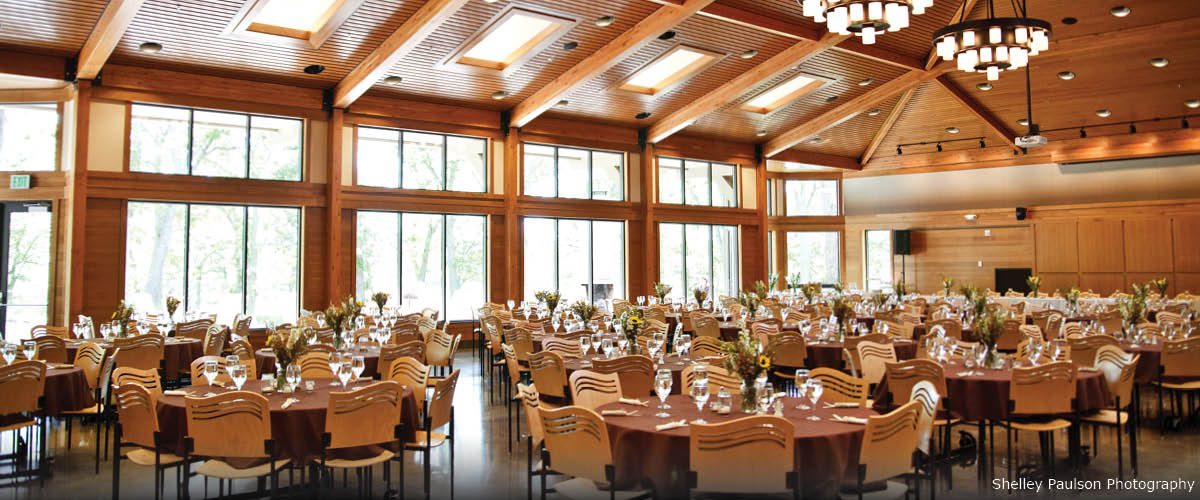 The inside of a great hall with vaulted wood ceilings, large chandeliers and a wall of windows. The room is set with tables and chairs for a wedding.