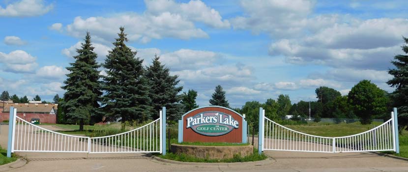 A sign at a gated entrance reads "Parkers Lake Golf Center." There are tall pine trees behind and a blue sky dotted with puffy clouds.