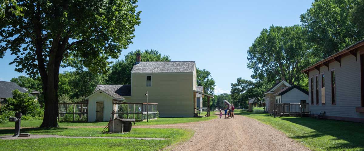 A few people walk down a dirt road that is lined with historic buildings.