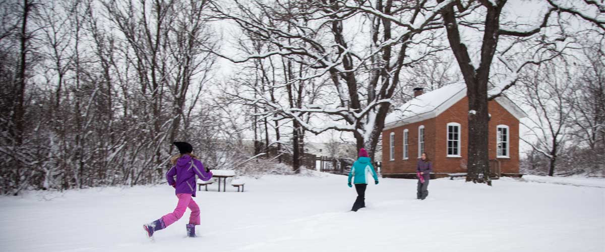 Two children run through the snow. A brick historic building is in the background.