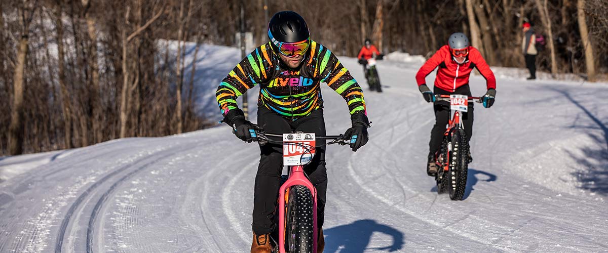 Fat bike riders compete in a race on a snowy trail through the woods.