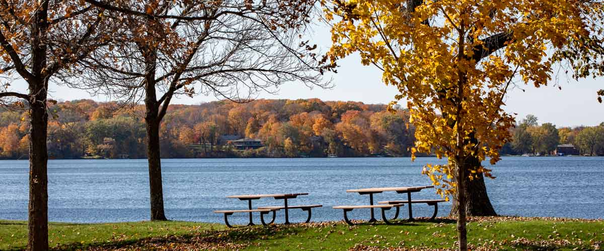 Two picnic tables in a lawn overlook a lake in the fall.