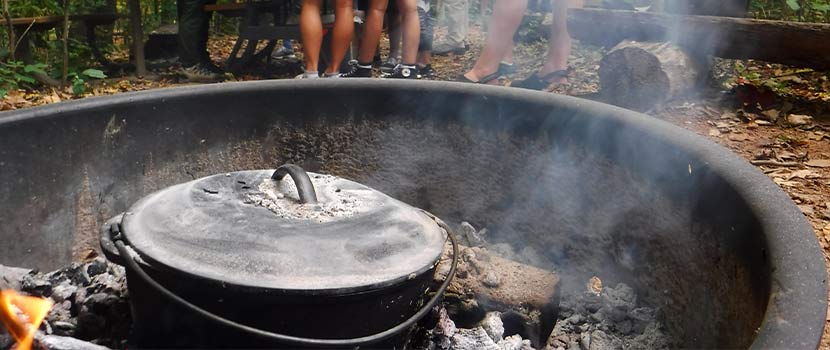 A dutch oven sits in a fire ring at a campsite.