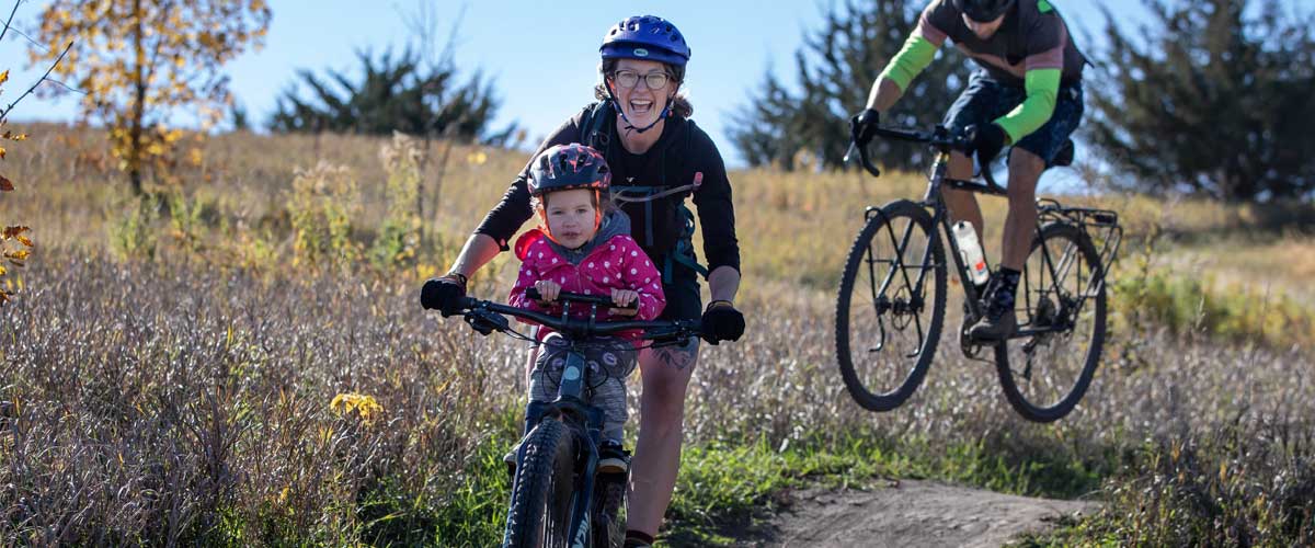 A woman and her daughter smile as the ride a mountain bike. Behind them, a man catches air on his mountain bike.