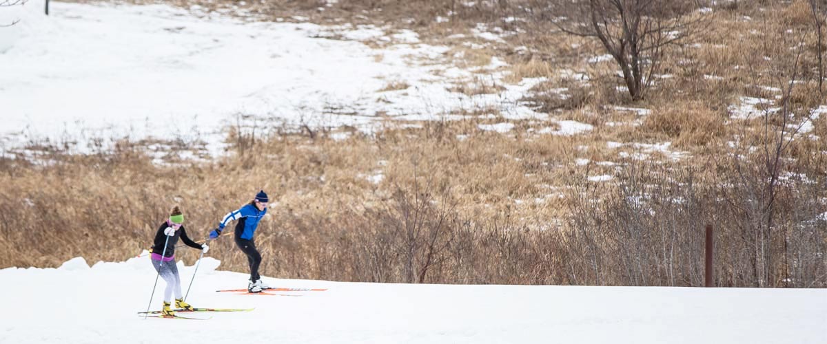 Two people cross-country ski on a snow-covered trail.