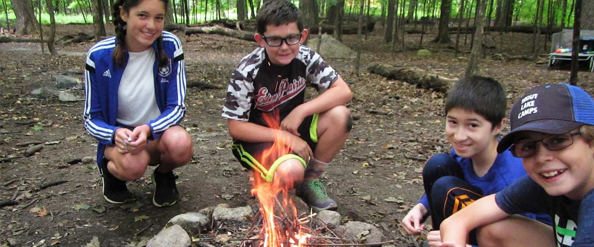Four kids smile as they gather around a fire at the Baker Outdoor Learning Center.