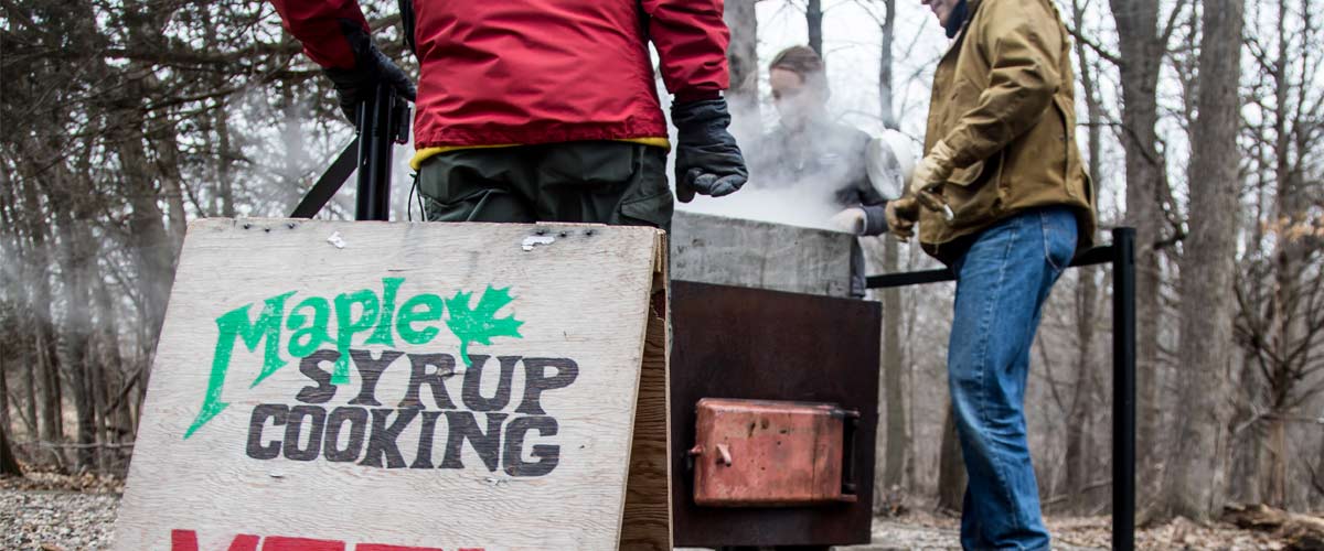 A plywood sign reads "Maple Syrup Cooking." Behind it a few people gather around as sap is boiled into syrup.