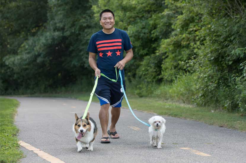 A man smiles as he walks two dogs on leashes down a paved trail.