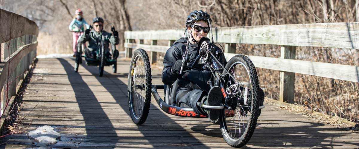 A person crosses a wooden bridge on a handcycle. Another handcyclist is close behind.