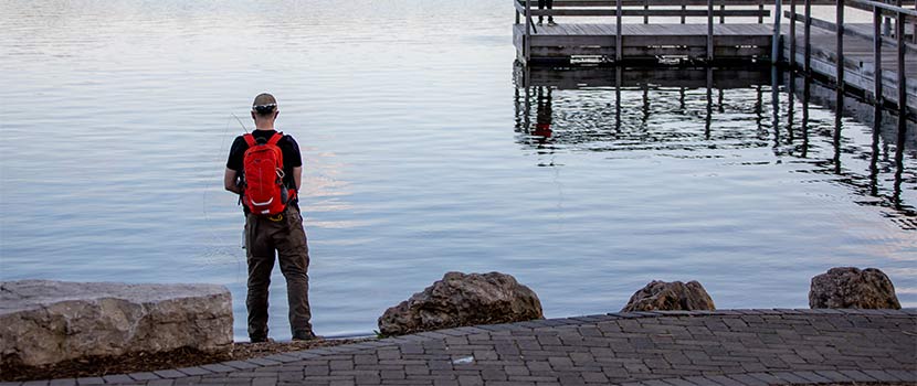 a person wearing a red backpack fishes on the shore of Silver Lake. A fishing pier is to their right.