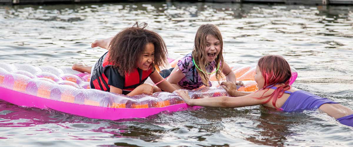 Three girls laugh as they swim with a large floating raft. 