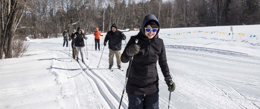 A woman wearing sunglasses smiles as she leads a group of people cross-country skiing on the snow.
