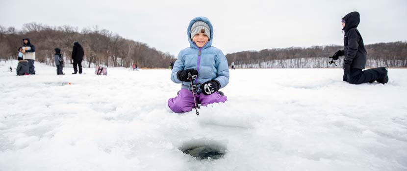 A child holds a fishing pole over a hole in the ice, bundled in winter clothing and smiling.