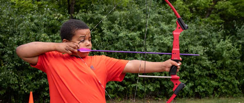 A boy wearing an orange shirt pulls back a purple arrow in a red and black bow.