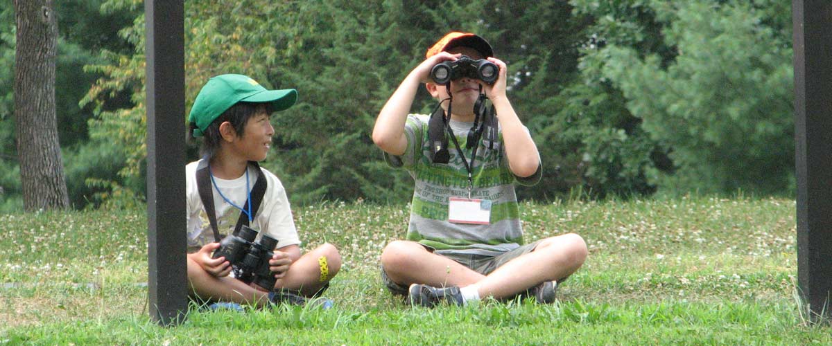 Two children sit on the grass. One looks through binoculars. The other is wearing binoculars around his neck.