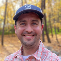 Josh, wearing a plaid shirt and navy baseball cap, smiles. Out of focus in the background are trees with yellow leaves.