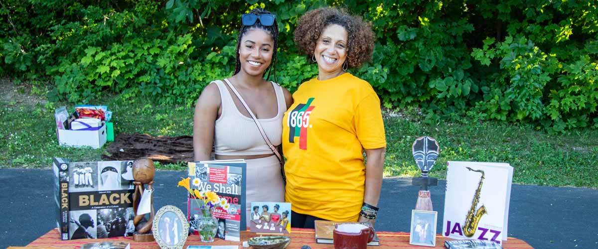 Two Black women stand behind a booth that offers information about Juneteenth, with books titled "Black," "We Shall Overcome" and "Jazz." 