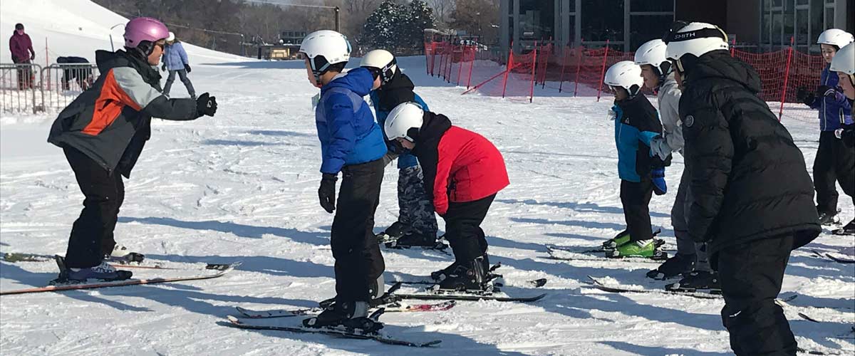 At the bottom of a ski hill, an instructor teaches kids wearing helmets and skis.