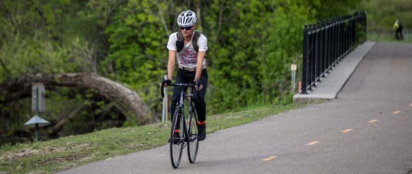 A person wearing a bike helmet bikes on a regional trail. Green foliage and a bridge are in the background.