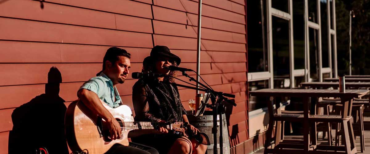 A guitarist and a vocalist sit behind microphones outside of the Hyland Hills Chalet.
