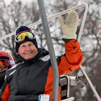 Roger smiles and waves to the camera with a mittened hand, while riding a ski lift. Ski goggles sit atop his head.
