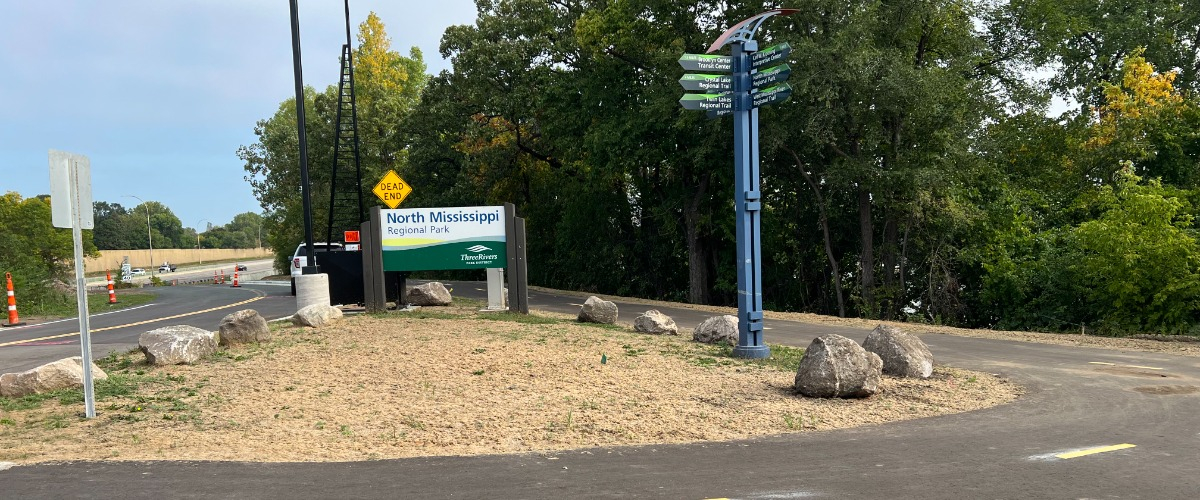 The North Mississippi Regional Park entrance including a paved trail, road, directional sign and trees and blue sky in the background