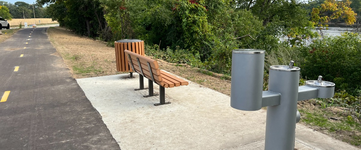 a bench, drinking fountain and garbage can along a paved trail with trees and the Mississippi River in the background