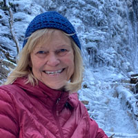 Bev, wearing a winter hat and coat, smiles at the camera with snow-covered trees in the background.