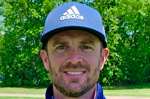 A man in a navy Adidas hat smiles for the camera in front of a golf course. 
